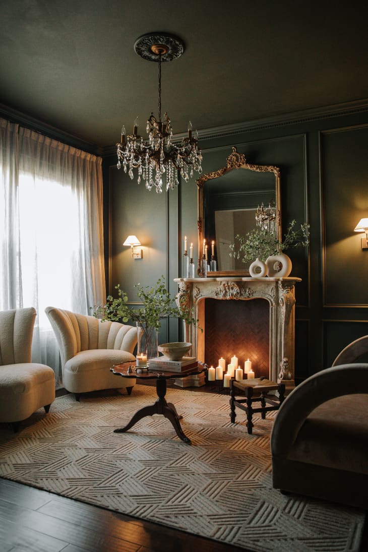 Living room with dark green walls, chandelier, ornate fireplace with candles, natural textile rug, European style furnishings