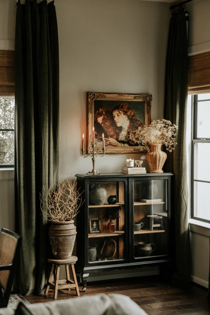 windows with brown shades and floor to ceiling green curtains, European painting in gold frame in corner over antique black cabinet
