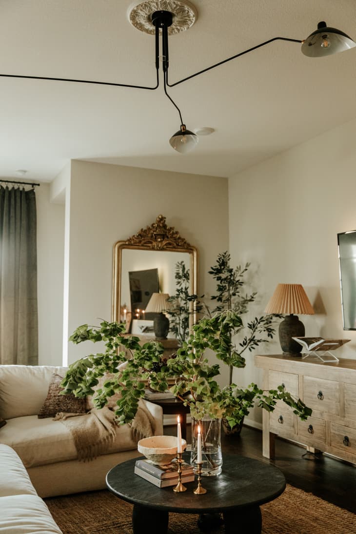 Corner of living space with neutral sectional sofa, round coffee table, large eucalyptus branches in vase, antique dresser/credenza with lamp, large wall mirror with baroque gold trim