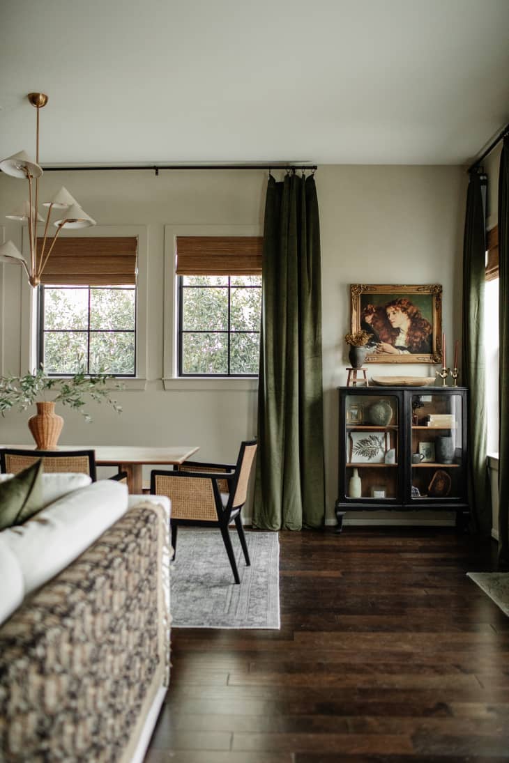View into dining area. Wood dining table with rattan and black chairs, windows with brown shades and floor to ceiling green curtains, European painting in gold frame in corner over antique black cabinet
