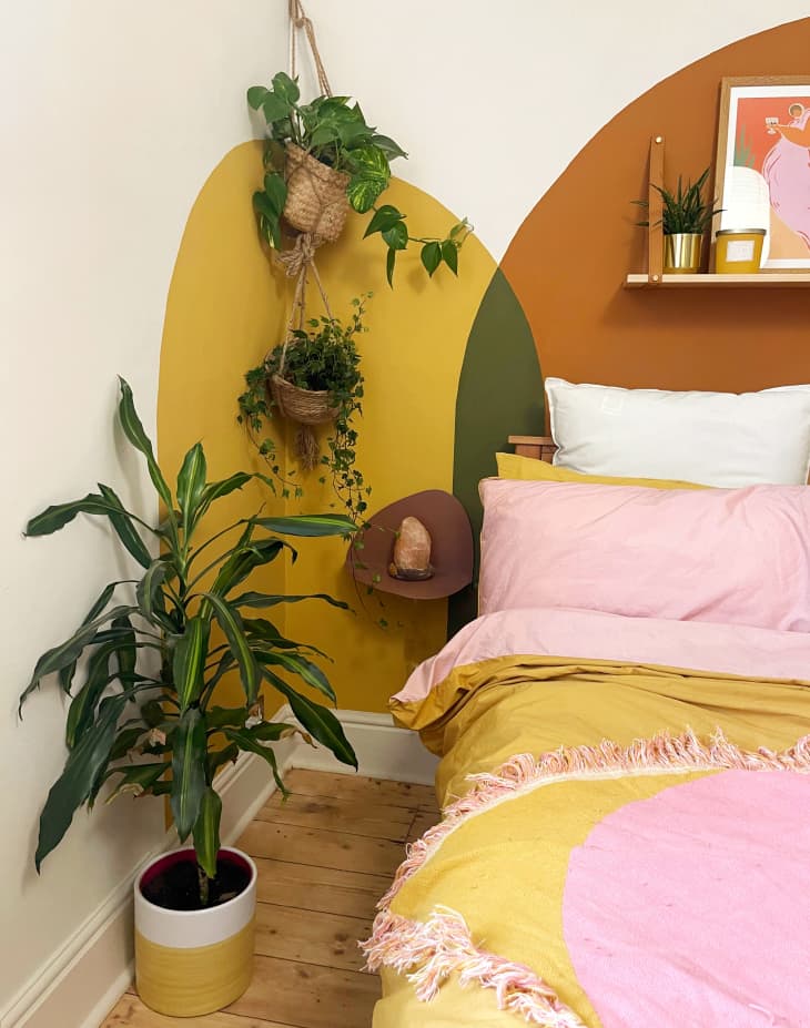 bedroom with rust, peach, and ochre painted shapes, plants, pink and yellow bedding