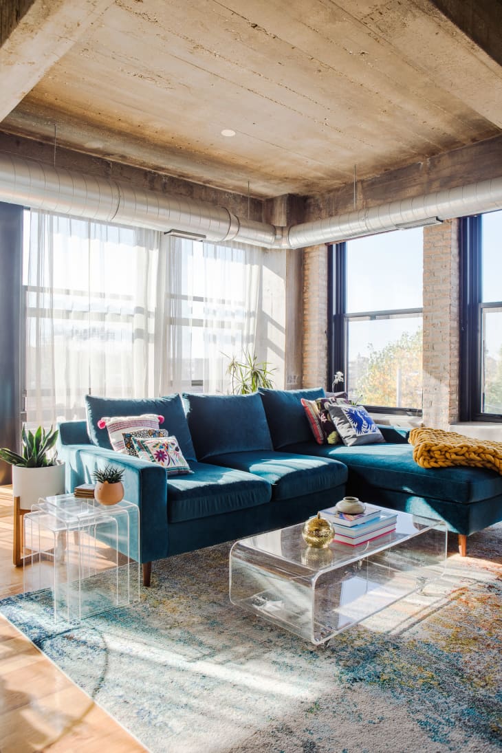 Living room with high ceiling, large windows, blue sofa with colorful throw pillows, lucite coffee table and side nesting tables