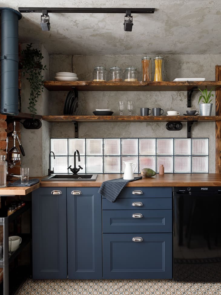 Kitchen with dark blue cabinets, glass block backsplash, open wood shelving, patterned tile floor, exposed brick wall corners