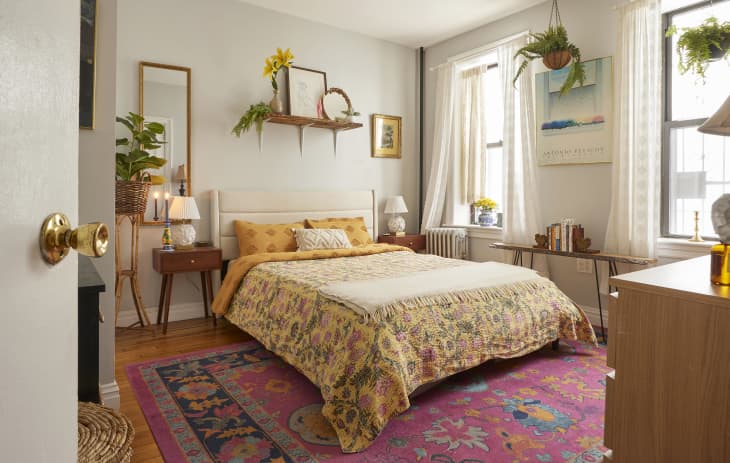 Plant filled bedroom with neatly made bed topped with pale yellow quilt. Colorful pink rug lines the floor.