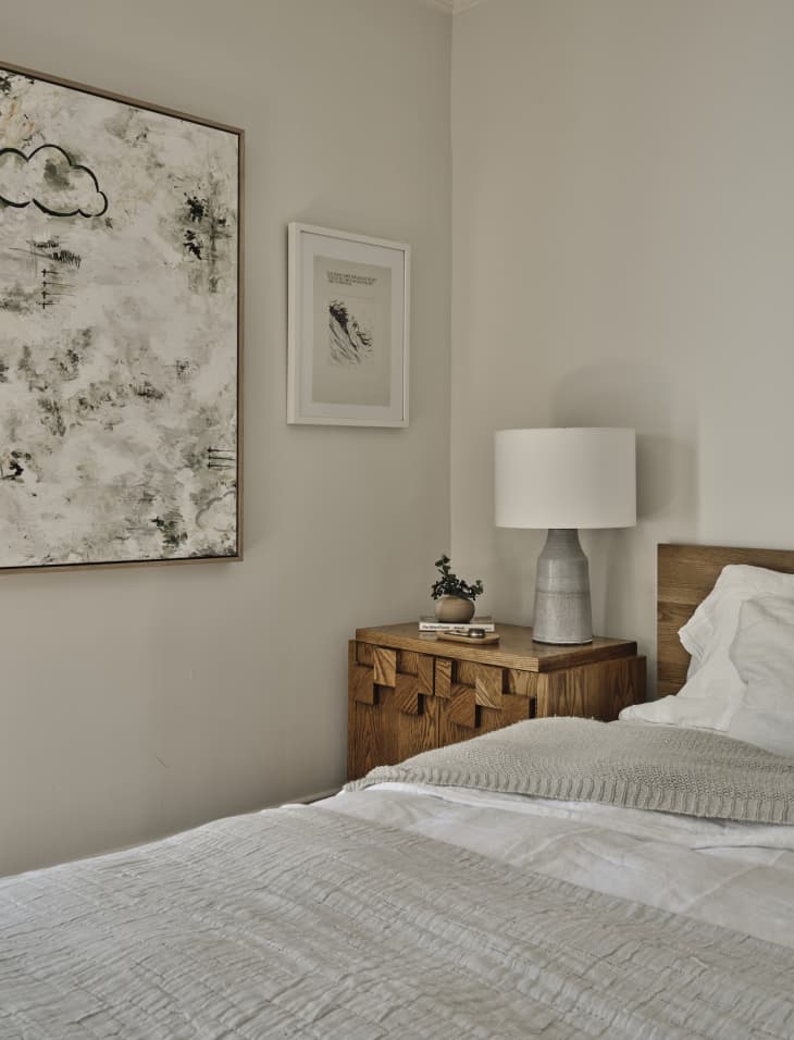 bedroom with pale gray walls, bed with pale gray and white bedding, wood headboard and nightstand with gray lamp with white shade. Framed art on wall