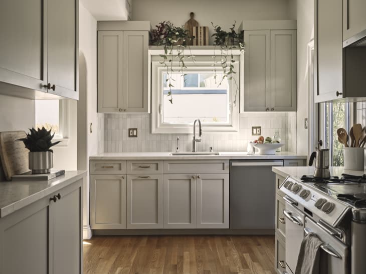 kitchen with wood floors, pale gray cabinets, pale gray vertical tile backsplash, window over sink with shelf above with plants, vintage oven/stove
