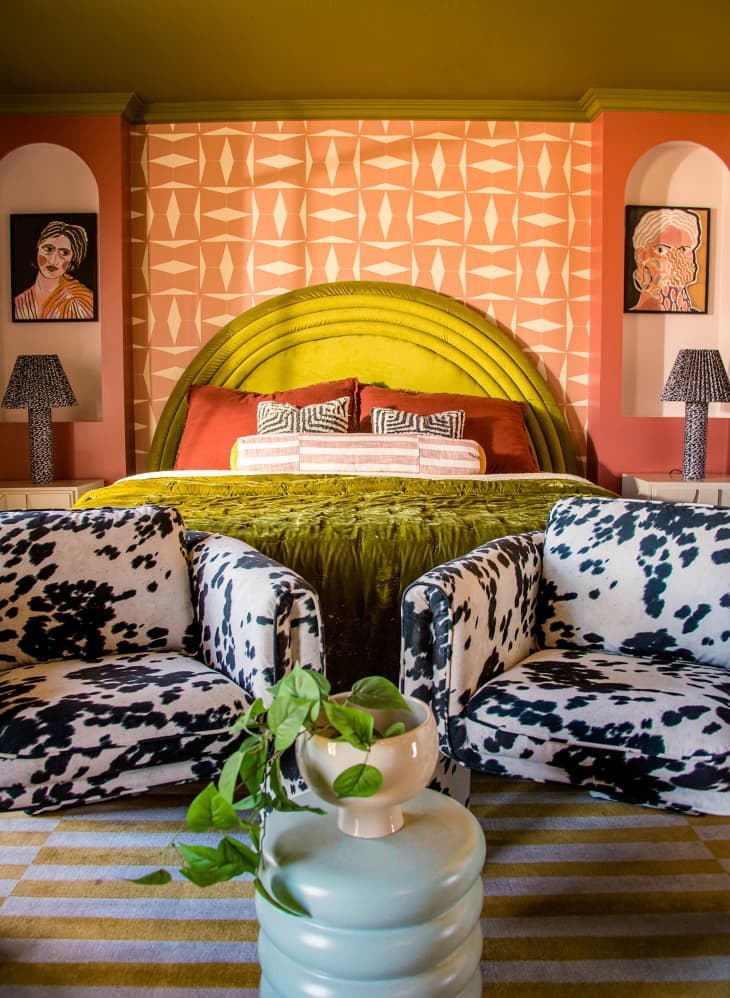 Colorful bedroom with orange and white graphic motif wall paper on accent wall. Chartreuse green upholstered headboard with two cow print arm chairs at the foot of the bed.
