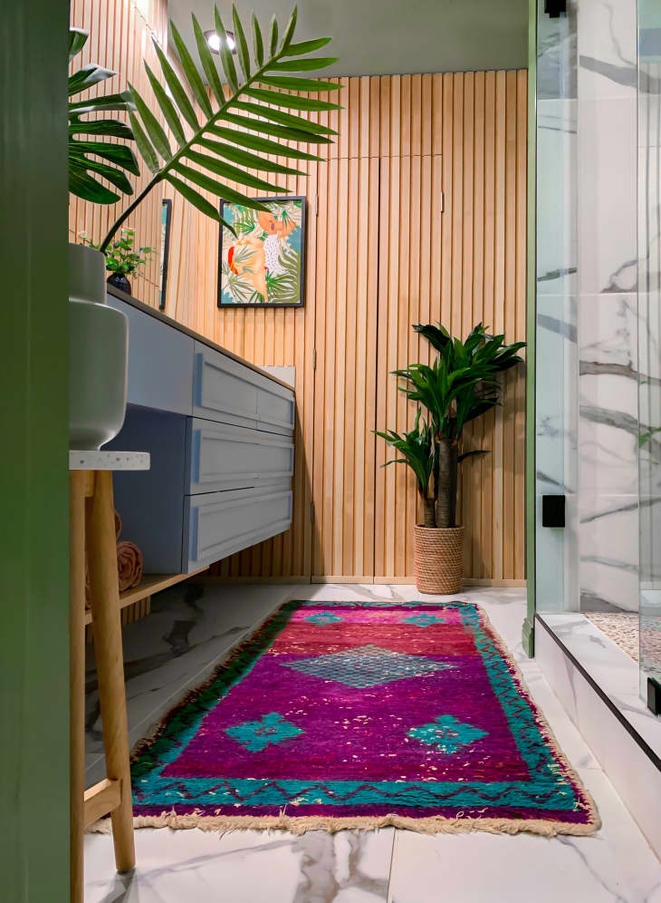 Plant filled bathroom with fluted wooden paneled walls and a blue vanity. Colorful rug lines the floor.