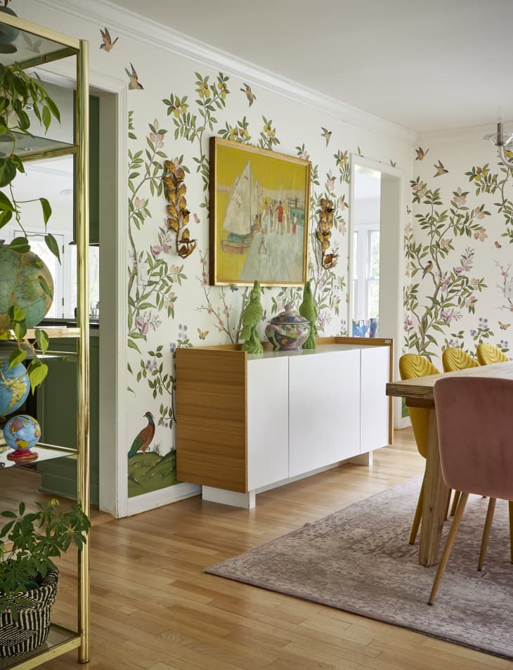 Painting mounted above white credenza in dining room with whimsical botanical wallpaper.