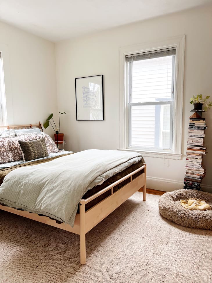 bedroom with pale neutral walls, light wood bed frame and striped linen duvet, patterned throw pillows. Pet bed on floor next to stack of books with a plant on top