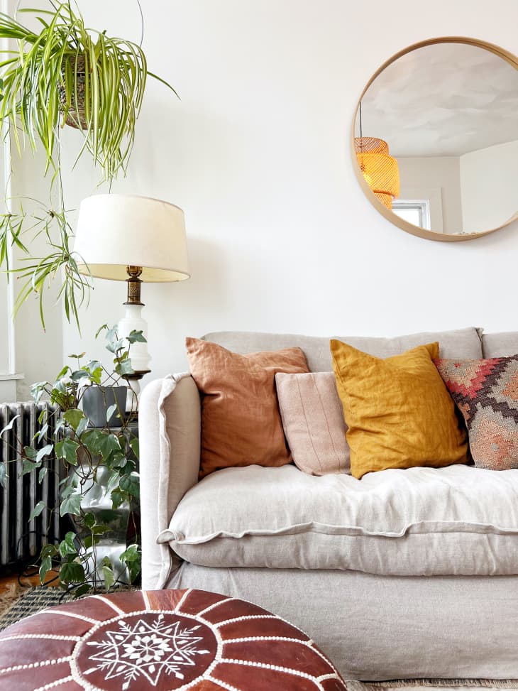 detail of sofa with cozy throw pillows, hanging plants, round mirror on wall, moroccan style leather pouf