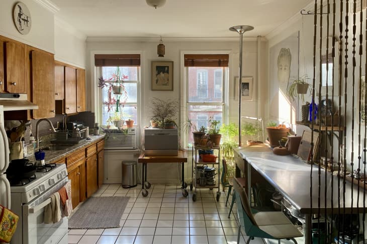 kitchen with 2 windows, wood cabinets, long table against wall with dining chairs, white tiled floor, rolling carts with plants, mini dishwasher