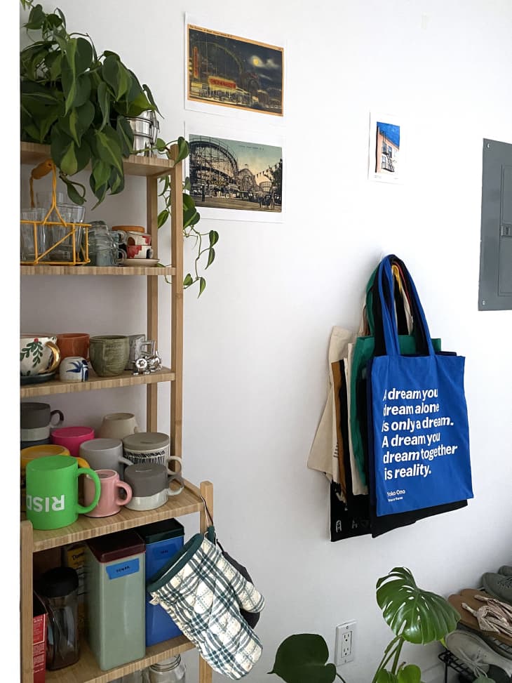detail of wall in entryway with wood shelves with colorful mugs, plants. Tote bags hanging on wall hook, plants on floor, white walls