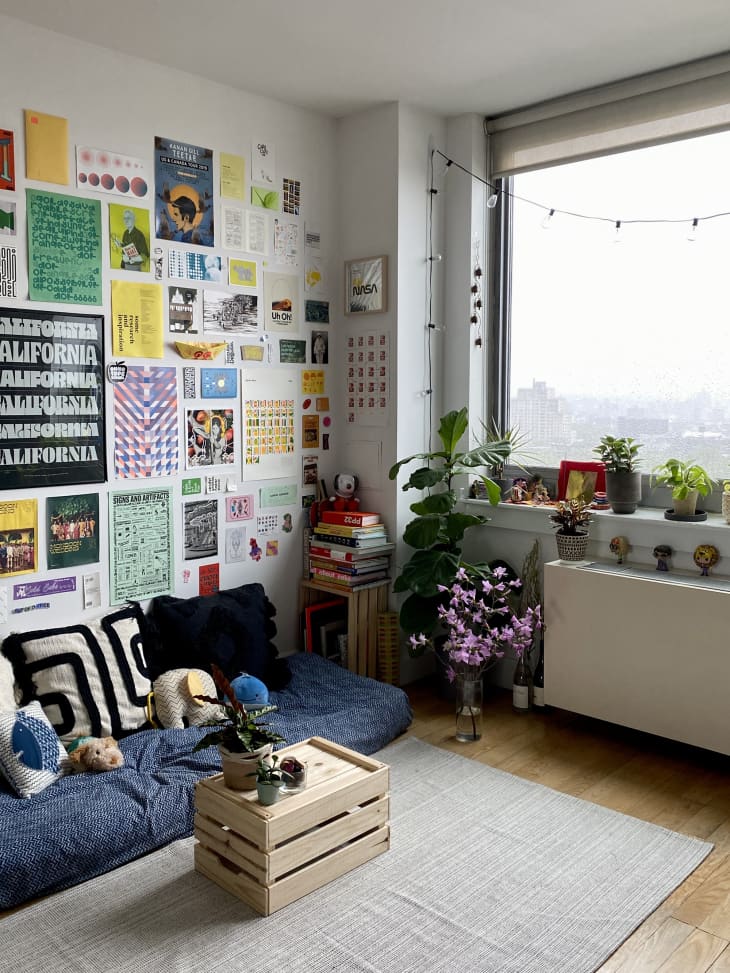view from entryway into living space in brooklyn apartment: cushioned seating on floor with navy blue cover, black white and navy throw pillows. unframed art gallery wall on wall behind. crate as coffee table and crate with stack of books. Plants on floor and radiator, large window, white walls, pale neutral area rug
