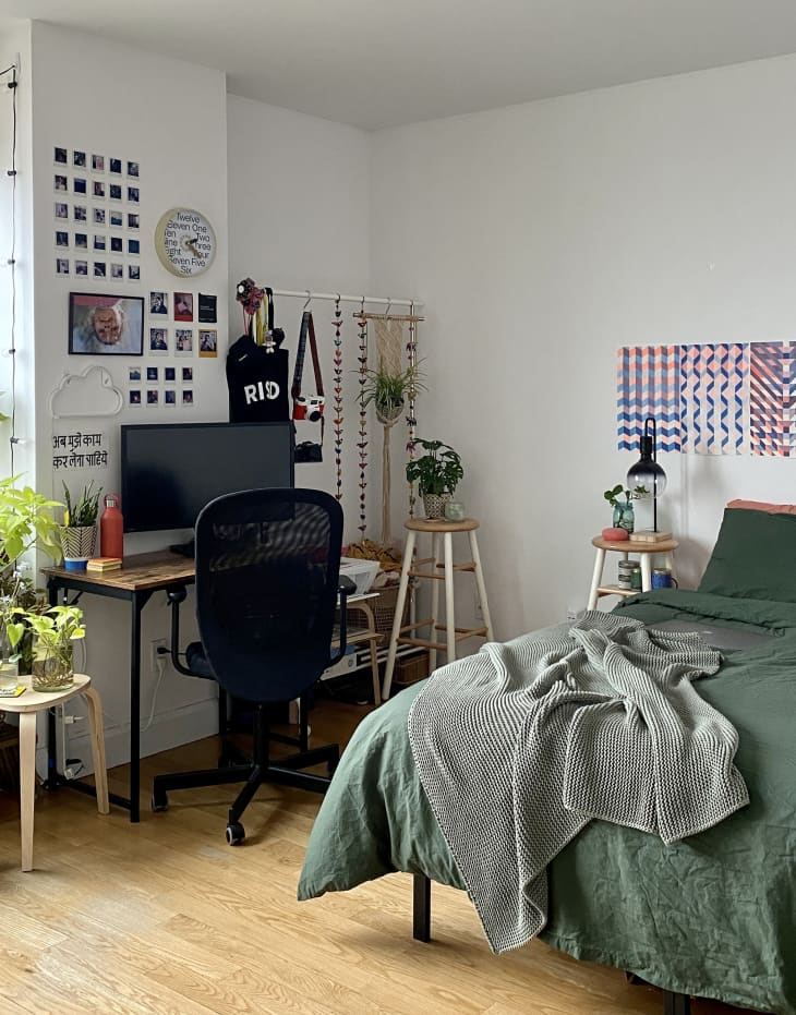 bedroom and workspace area in apartment with bed with green linens, lots of unframed art on walls, wood desk with black metal legs and black office chair, small table with plants, white walls