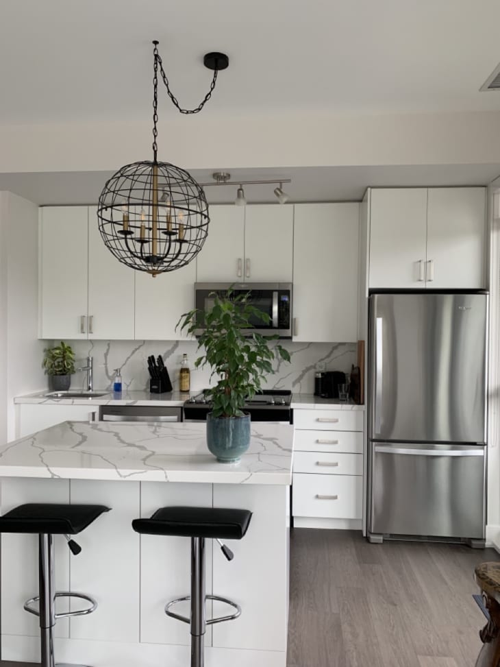 White modern kitchen with white counters and cabinets/drawers, stainless steel fridge, round metal "cage" over candelabra as light fixture, black barstools