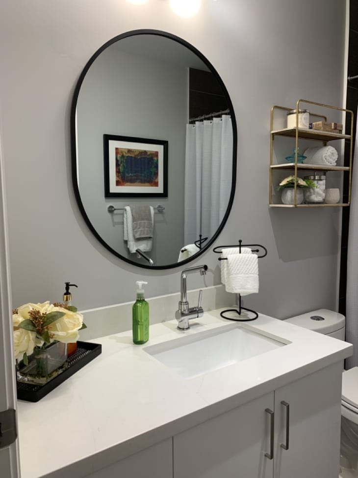 Bathroom with gray walls, oval mirror with black frame over sink, white counter and cabinets, black and gold accents