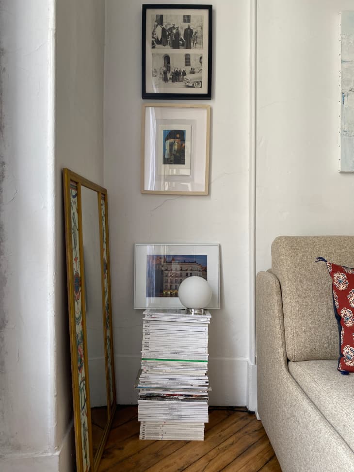 Stack of magazines on apartment floor with small lamp on top. Art prints on wall above magazine stack.