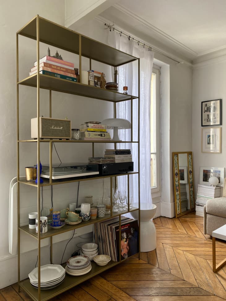 Gold bookshelf serves as link between kitchen and living room. Bookshelf is decorated with books, cockery and a speaker in Parisian apartment.