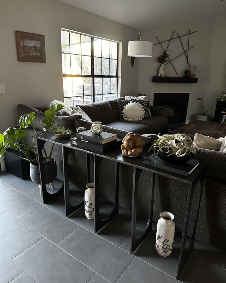 Living room with black console table with vases and art objects, dark gray sofa with black and white pillows, plants, large paned window, gray tiled floor