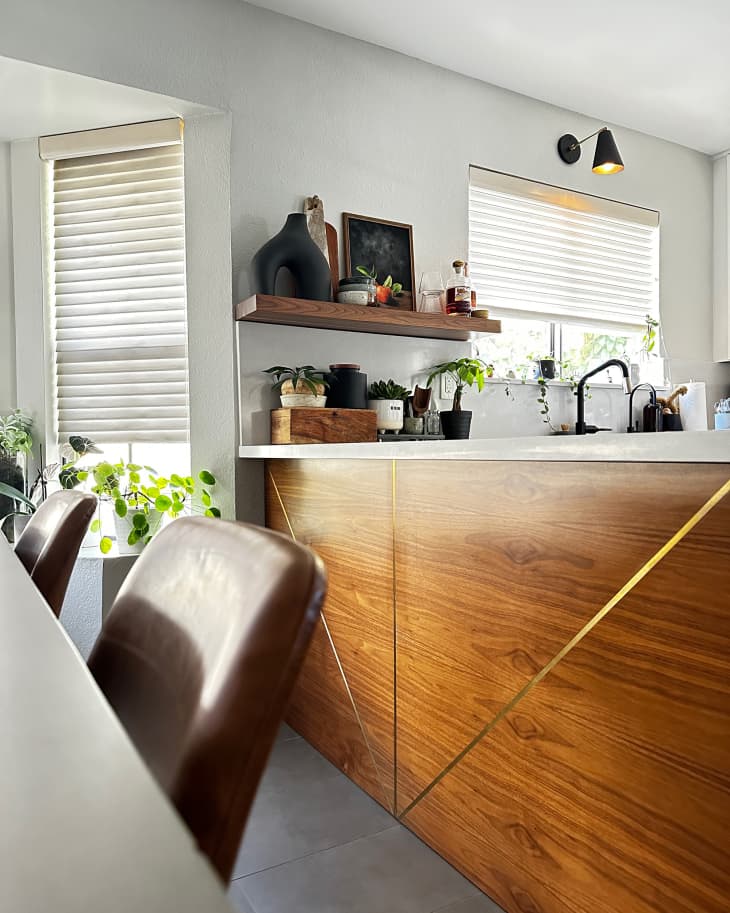 kitchen with wood counter block with gold detail, white countertop, floating wood shelves, part of dining table and chairs showing