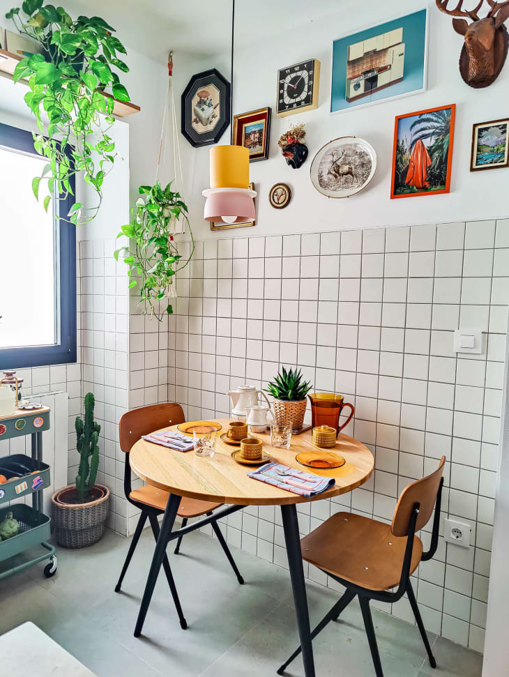 Breakfast/dining nook with small round table, 2 wood and metal chairs. Table is set with orange and terra cotta dishware. White tile wall and gallery wall above. To the left there's a window and hanging plants. Colorful small pendant light