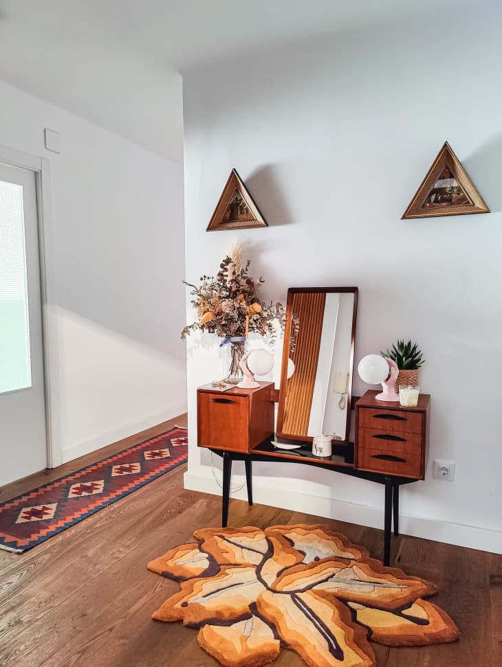 corner of room with white walls, small table with drawers and a rectangular mirror, small flower-shaped area rug in warm hues, 2 triangular-framed paintings on wall above. View into hall with patterned runner in warm hues
