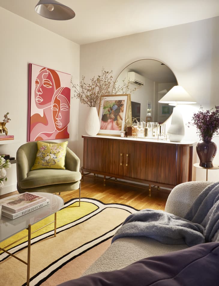 wood credenza in corner of living room with large round mirror above. White lamp and large white vase with dried flowers on top, Art and other objects. Wood floors, white walls, we can see a velvet green chair, striped rug, and  part of a  coffee table