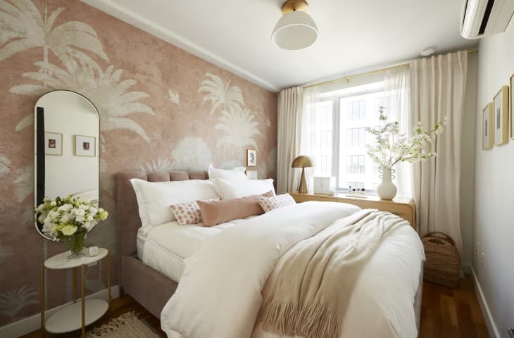 Bedroom with velvet quilted headboard and bed frame, white linens, blush velvet pillow, oval mirror on wall over small white table with vase of white flowers. Wall has palm tree themed wallpaper in blush and cream. Large window with off white curtains