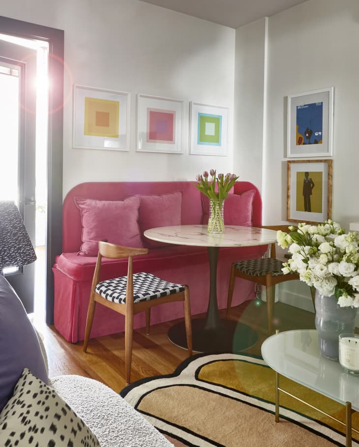 Living room with pink velvet loveseat with small round table, 2 extra chairs for dining, framed artwork on wall above. White oval coffee table in front of textured gray loveseat. white shelves under a wall mounted TV with books, a gold vase. Vases of flowers on both tables