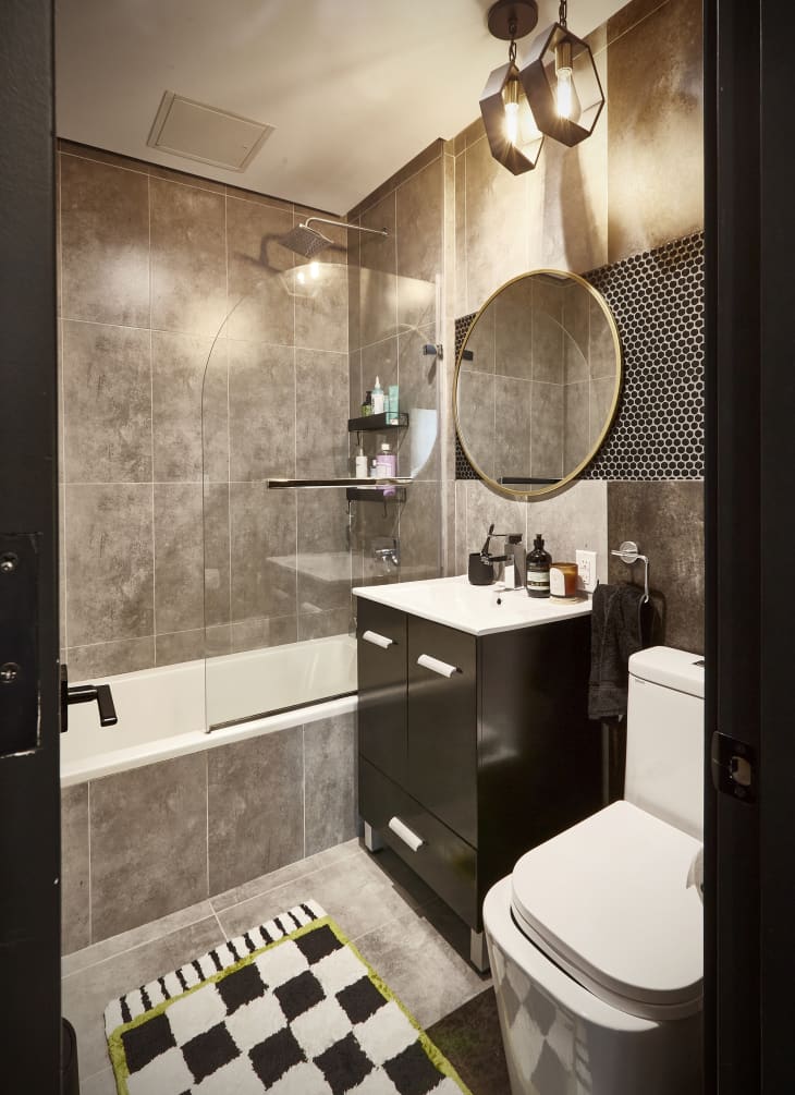 Bathroom with large brown tile walls, shower, and floor. Black and white checkered rug, round brass trim mirror over sink. 2 hanging lights over sink, part of wall behind mirror is small round tiles