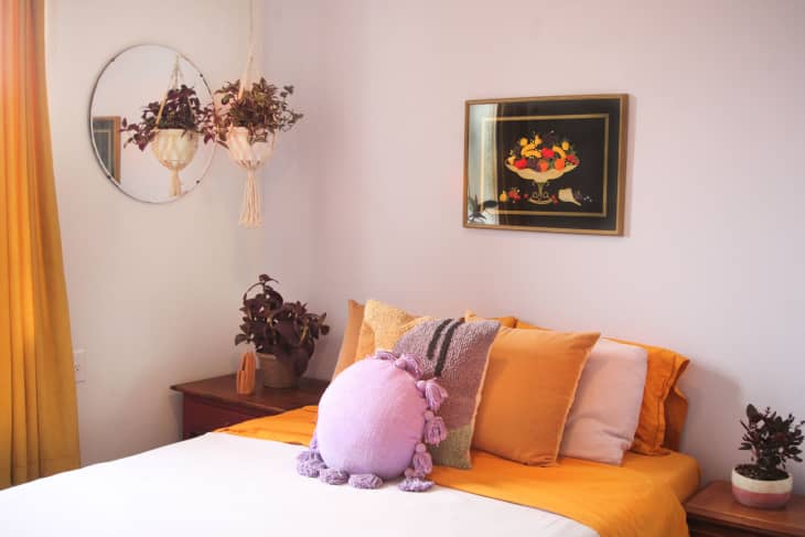 Bedroom with pale mauve walls, bed with lilac and pale orange bedding, 2 end tables with plants, one plant hanging in macrame planter, framed art above bed