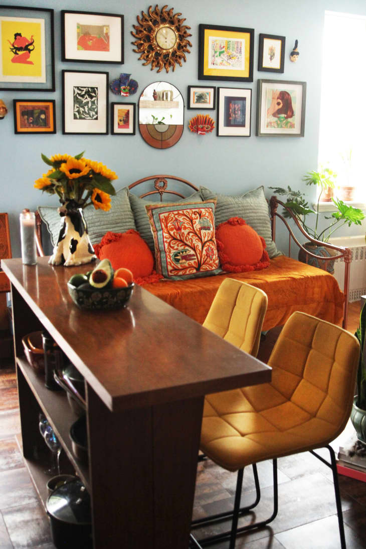 view of dining and living area in small apartment. Dining "table" is a console table with shelves, 2 cushioned chairs pulled up. There's a vase of sunflowers on top. In the background is the living room sofa/daybed with an orange velvet throw/cover and pale greenish blue and orange throw pillows. Gallery wall on blue wall above sofa