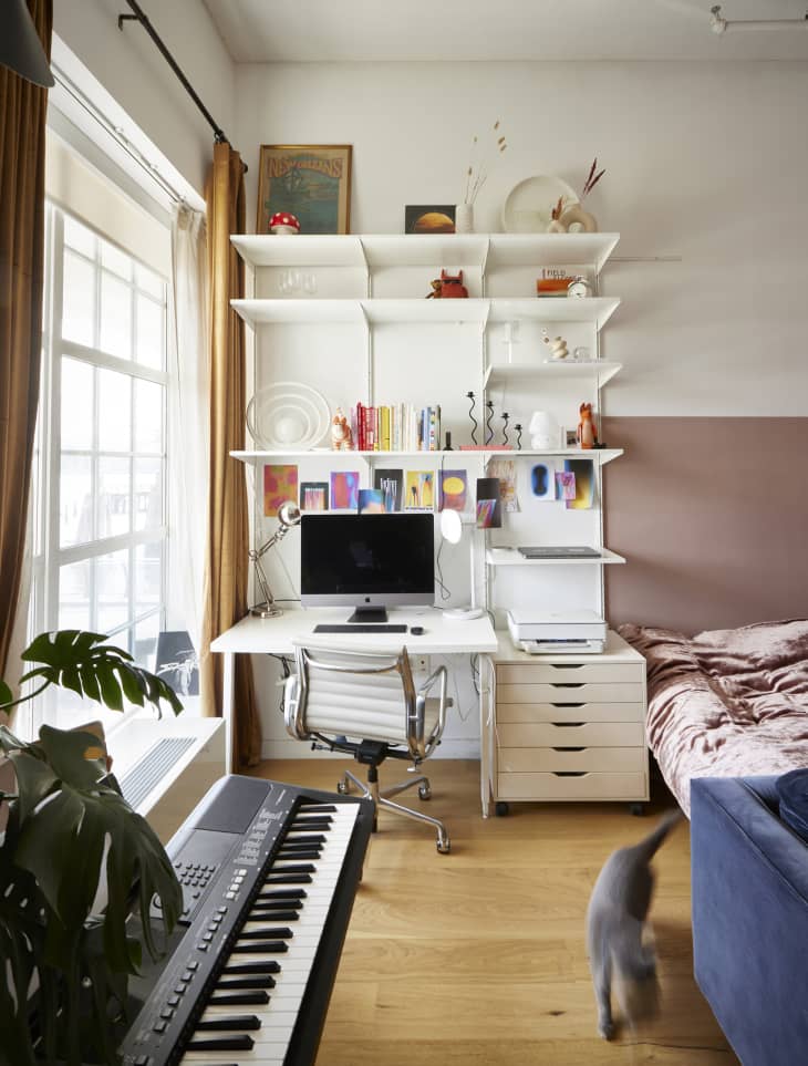 desk/office home workspace with computer, gray task chair, white shelves to ceiling. Bed to the right with muted rose wall, gray cat and piano keyboard in foreground