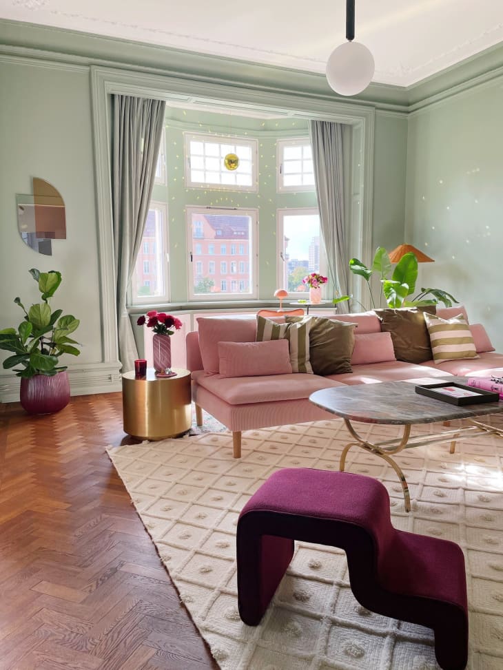 after photo of living room: living room with green walls, bay window with white trim, green curtains, gold disco ball in window, pink velvet sofa with velvet and striped throw pillows, textured diamond pattern rug, oval marble coffee table, small table and chair in window, vase of pink flowers, large potted plant on floor