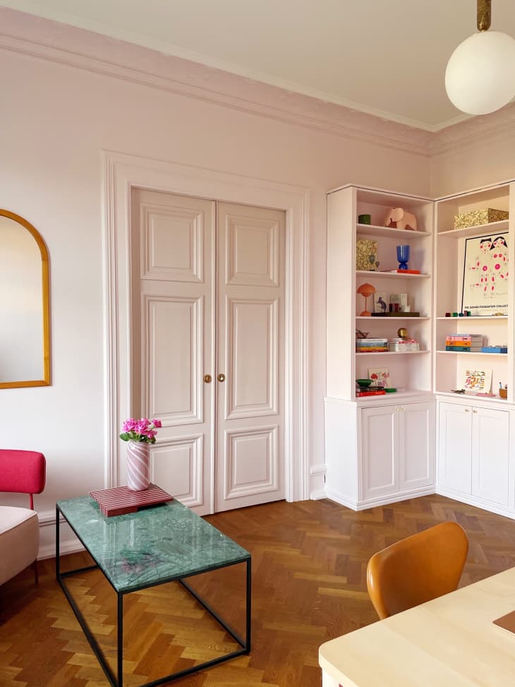after photo of studio: pale pink walls, pale pink shelves with decorative objects, pink doors, parquet wood floor, green stone coffee table with pink flowers in vase, arched gold frame mirror