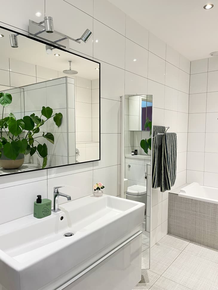 White bathroom with large tile walls, large black framed mirror over wide sink, potted plant reflected in mirror