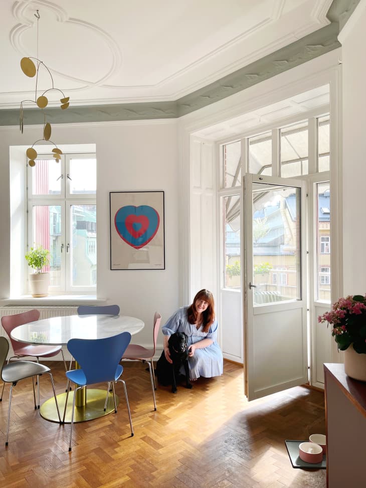 dining area with white walls, round glass dining table with different colored midcentury chairs, parquet wood floor, framed heart poster, window with potted plant on sill, doors open to balcony with table and chairs. Woman and black dog
