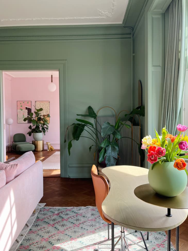part of living room with small curved table with one chair and pot of bright colored flowers. Green walls, part of pink velvet sofa visible. Another room viewed through door with pale pink walls, large potted plant