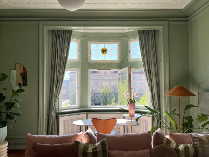living room with green walls, bay window with white trim, green curtains, gold disco ball in window, pink velvet sofa with velvet and striped throw pillows, textured diamond pattern rug, oval marble coffee table, small table and chair in window, vase of pink flowers, large potted plant on floor