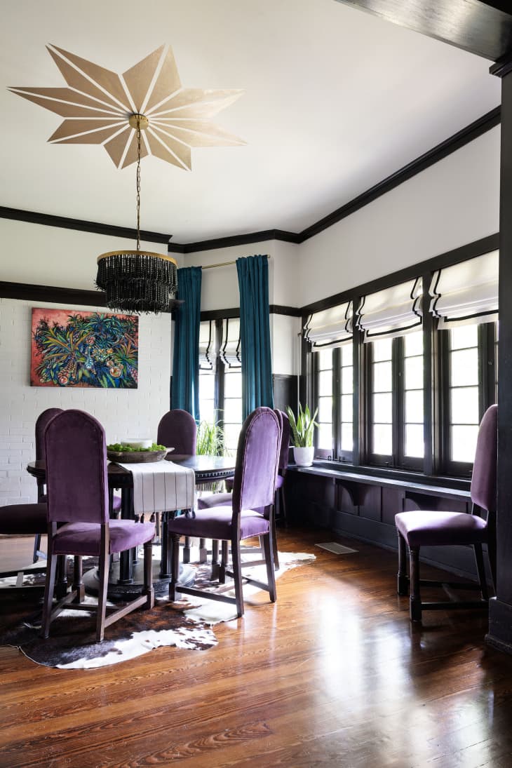Dining area with black table with purple velvet dining chairs, white walls with black trim, black and white window shade, wood floors, black fringed pendant light hanging from snowflake/star shaped painted design on ceiling