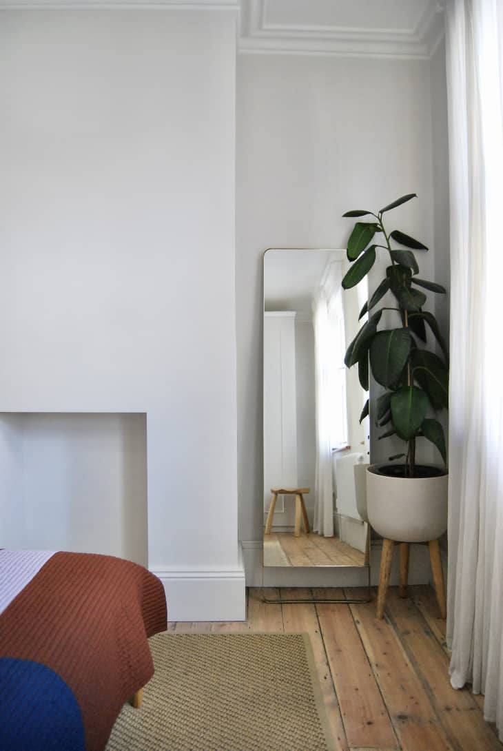 Large houseplant sits in front of standing mirror in corner of all white bedroom.