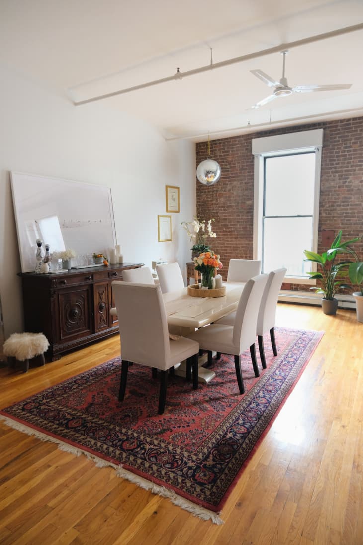 Dining room with white table and dining chairs with area rug, white walls, high ceilings, wood floors, white ceiling fan, credenza with large mirror, one exposed brick wall in background