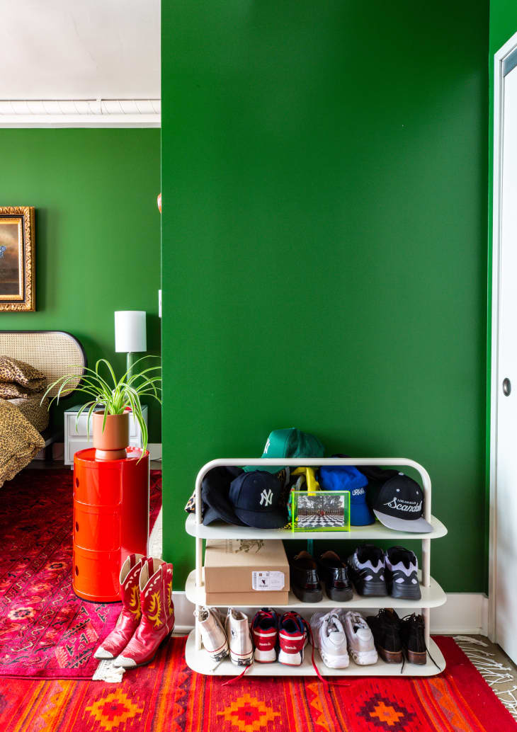 corner of bedroom with red patterned rug, shoe rack with hats and shoes, red small round drawer cabinet with plant, red cowboy boots on floor, green walls