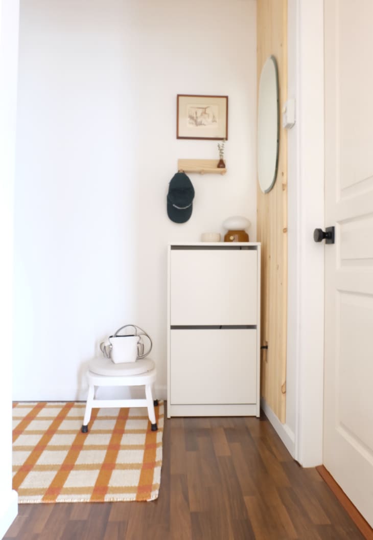 Corner in entryway of home with white walls, white cabinets, orange and cream flatweave rug, oval mirror, small floating shelf with hooks (hat hanging on one)