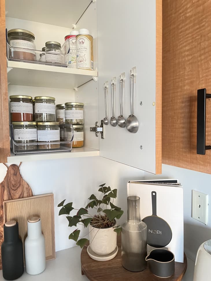 Open kitchen cabinet with jars of spices, measuring spoons hanging on door, cutting boards and containers on counter