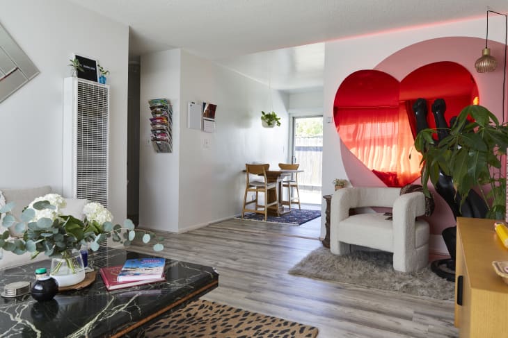 white living room with gray wood finish flooring, large red mirrored heart on wall, pale gray sofa and chair, black marble coffee table, and animal print rug