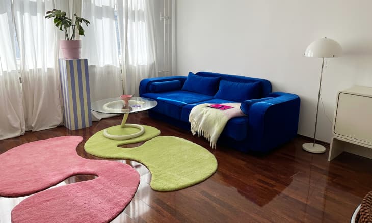 A white-walled living room with a blue couch with matching green and pink rugs.