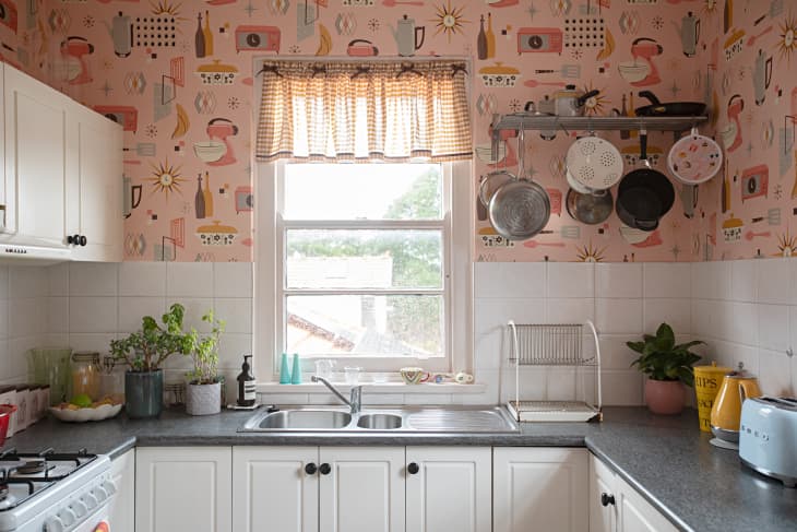 A kitchen with white cabinets, grey counter, white back splash and pink vintage appliance wallpaper.