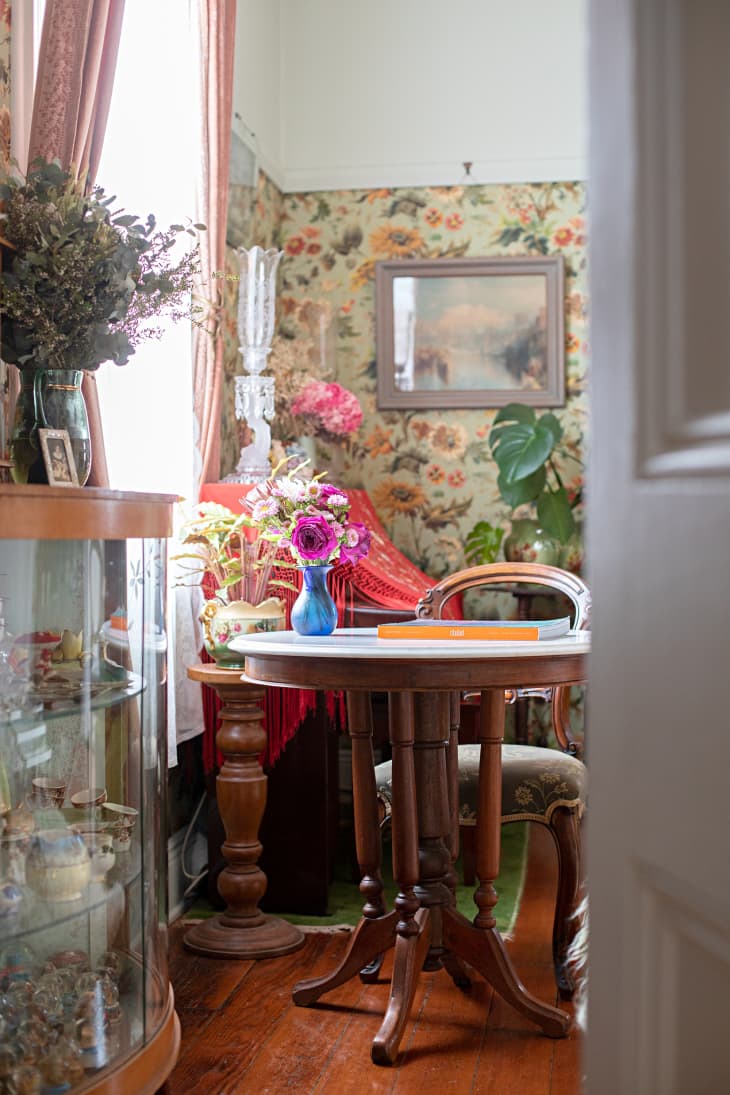 A living room with floral wallpaper walls with a table with vased flowers, books and a chair.